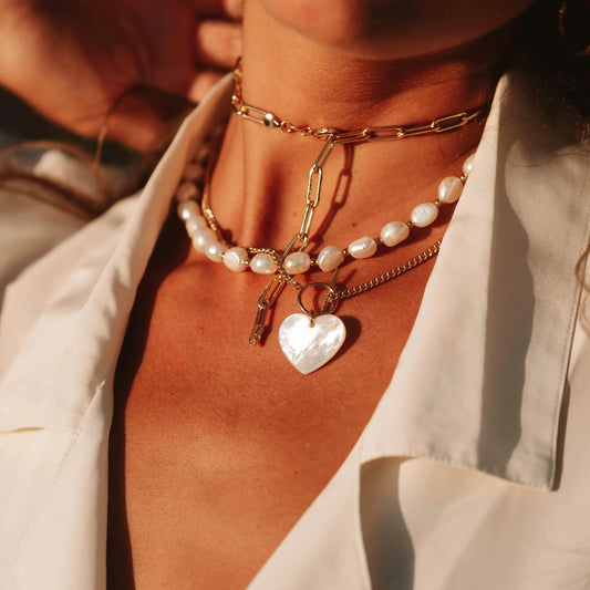 Close-up of a woman's neck adorned with layered gold and pearl necklaces, featuring a heart-shaped mother-of-pearl pendant, captured in warm sunlight.