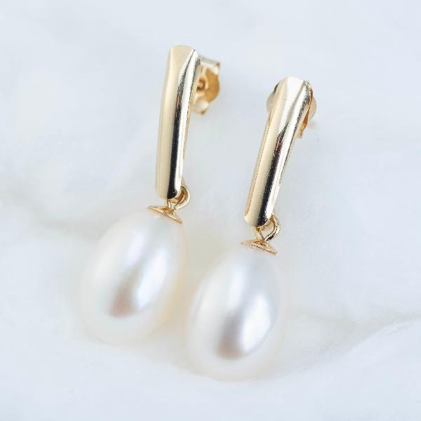 A pair of 14K Yellow Gold Freshwater Pearl Drop Earrings by NY Gold & Silver Trading Inc. dangle elegantly against a soft white fabric background, exuding sophistication with their sleek, modern design that captures the essence of classic pearl jewelry.