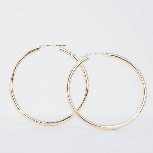 Two 14k yellow large gold hoop earrings (3mm)  from the Goddess Jewelry Collection  shine against a plain white background, showcasing their simple, polished design with a smooth surface reflecting light.