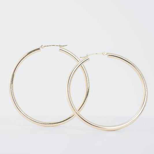Two 14k yellow large gold hoop earrings (3mm)  from the Goddess Jewelry Collection  shine against a plain white background, showcasing their simple, polished design with a smooth surface reflecting light.