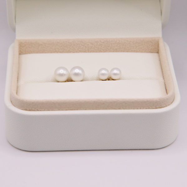 A white jewelry box with Freshwater Pearl Stud Earrings Duo Gift Set displays two pairs of pearl earrings on a beige cushion, arranged in two rows with one pair slightly larger, embodying timeless beauty.