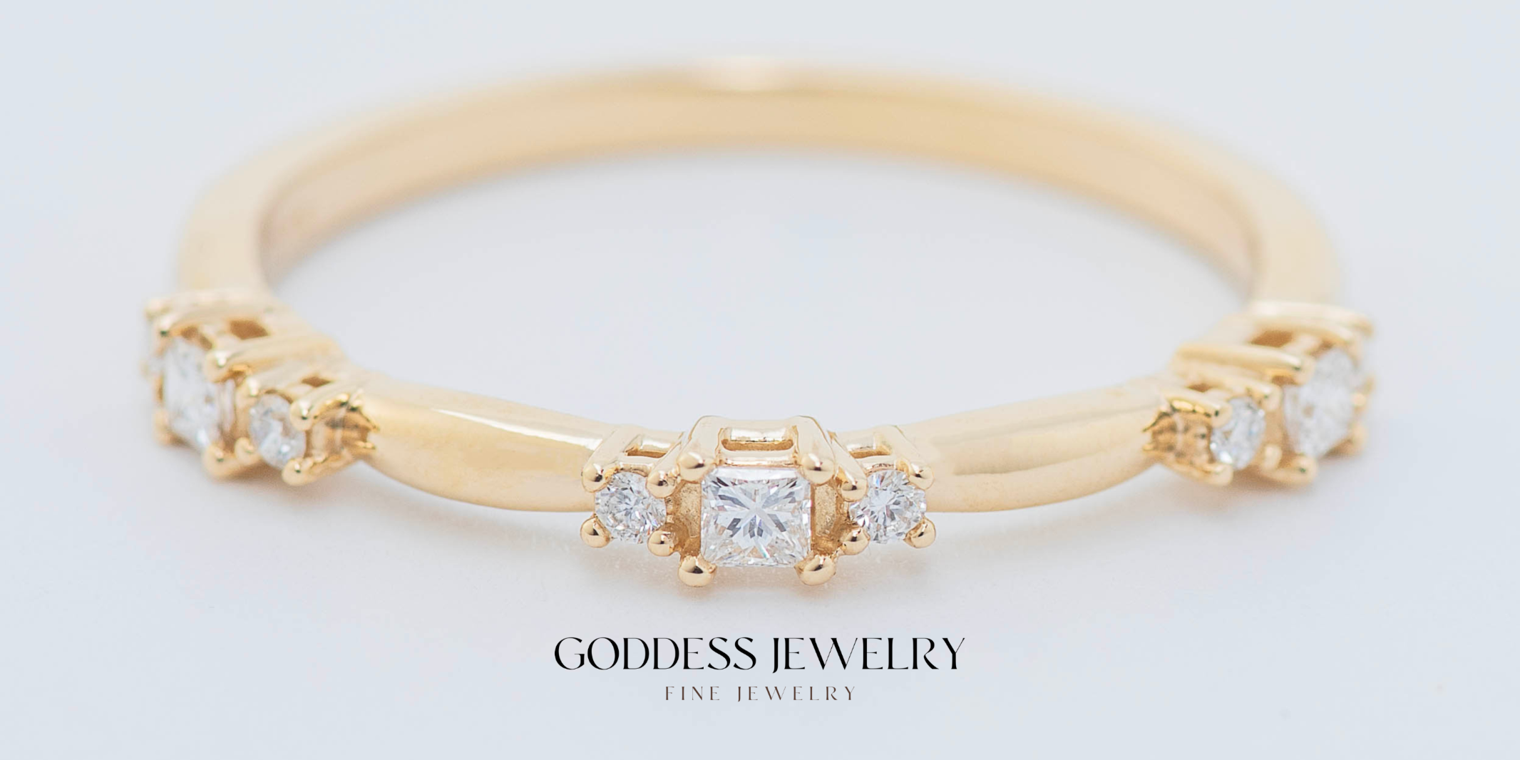 A gold ring adorned with a series of small diamond stones gleams against a white backdrop. Below the ring, elegant text reads, "Goddess Jewelry Fine Jewelry," showcasing timeless craftsmanship.