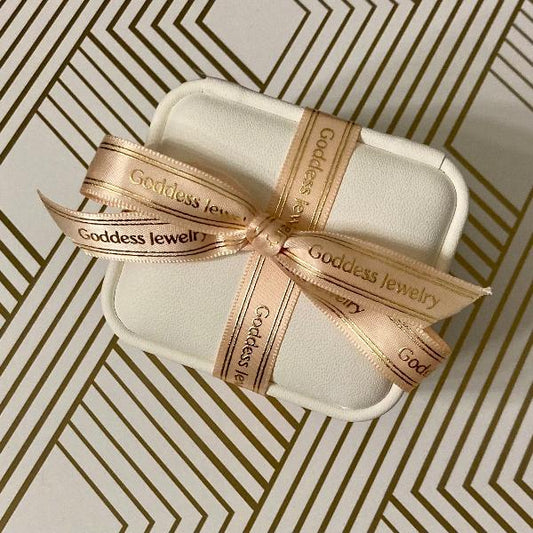 A white jewelry box, decorated with "Goddess Jewelry" in gold text and tied with a pink ribbon, lies stylishly on a gold and white geometric patterned background, suggesting the luxury of the 14k White Freshwater Pearl Stud Earrings Gift Set inside.