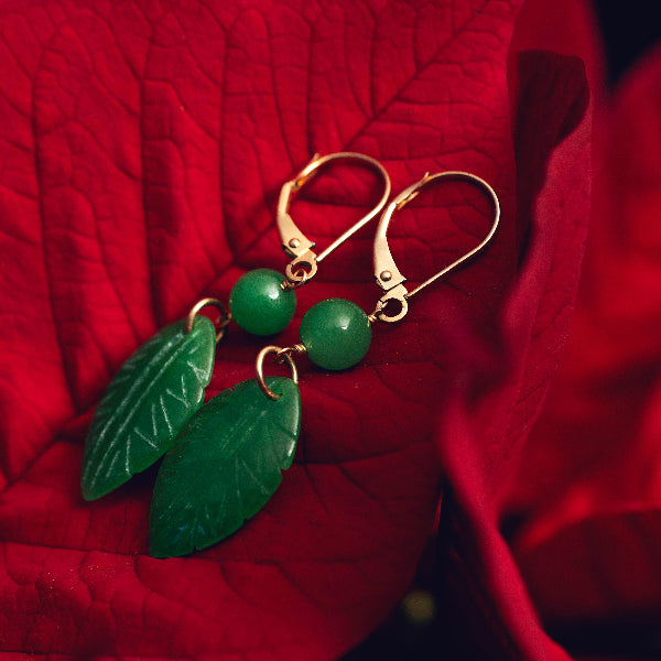 Elegant gold and green jade leaf earrings from the Goddess Jewelry Collection rest on vibrant red poinsettia petals, creating a striking contrast between the deep green gemstones and the bold crimson background. The intricate leaf carvings and polished jade beads add a natural, luxurious touch.