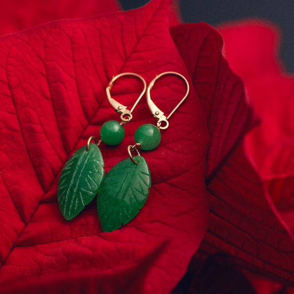 Elegant gold and green jade leaf earrings from the Goddess Jewelry Collection rest on vibrant red poinsettia petals, creating a striking contrast between the deep green gemstones and the bold crimson background. The intricate leaf carvings and polished jade beads add a natural, luxurious touch.