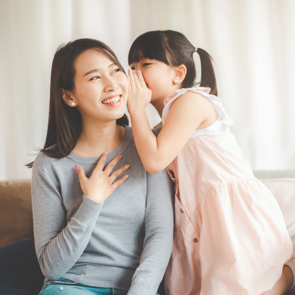 A young girl whispers to a smiling woman on a sofa, her light blue sweater glowing in the room. The girl's pink dress and ponytails bounce as she chatters about treasures like Freshwater Pearl Stud Earrings Duo Gift Set from the Goddess Jewelry Collection  , set against a softly blurred background.