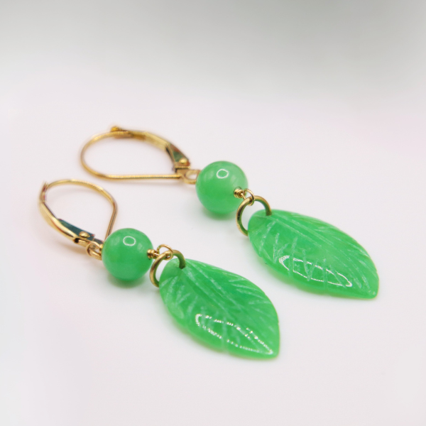 Elegant gold and green jade leaf earrings from the Goddess Jewelry Collection are displayed on a white marble background with delicate gold veining. The intricately carved jade leaves and polished green beads exude a natural, luxurious charm, enhanced by the refined gold-tone hardware.