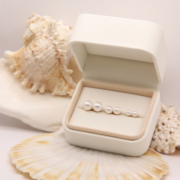 The 14k White 9mm Freshwater Pearl Stud Earrings from the Goddess Jewelry Collection are beautifully displayed in a row from large to small, with seashells elegantly arranged behind the display on a neutral background.
