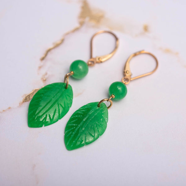 Elegant gold and green jade leaf earrings from the Goddess Jewelry Collection are displayed on a white marble background with delicate gold veining. The intricately carved jade leaves and polished green beads exude a natural, luxurious charm, enhanced by the refined gold-tone hardware.