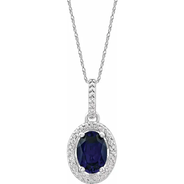 The Sterling Silver Lab Blue Sapphire Oval Necklace  from the Goddess Jewelry Collection features a delicate rhodium-plated chain and a pendant with a lab-grown blue sapphire surrounded by small clear crystals, all displayed against a white background.