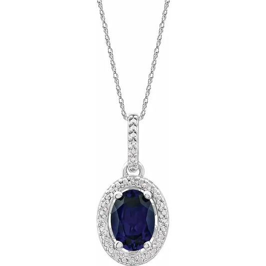 The Sterling Silver Lab Blue Sapphire Oval Necklace  from the Goddess Jewelry Collection features a delicate rhodium-plated chain and a pendant with a lab-grown blue sapphire surrounded by small clear crystals, all displayed against a white background.