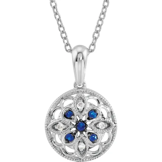 The Sterling Silver Blue Sapphire Filigree Necklace  from the Goddess Jewelry Collection features a round pendant with intricate filigree, adorned with lab-grown blue sapphires in a floral pattern and surrounded by clear stones for elegance, completed with a simple link chain.
