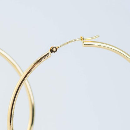 Close-up of a 14K yellow gold hoop earring on a light gray background. The polished finish and simple latch clasp of the 14k Yellow Gold Hoop Earrings 2mm exude timeless appeal. In the blurred background, another partial hoop adds depth to the scene.