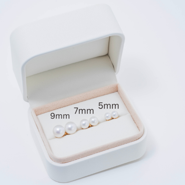 Displayed within an open jewelry box lined with soft beige fabric are three classic pairs of freshwater pearl stud earrings in various sizes, labeled 9mm, 7mm, and 5mm from left to right. The elegant arrangement showcases signature piece: the 14k White Freshwater Pearl Stud Earrings, ready to enhance any outfit with their timeless elegance.