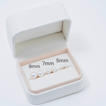 Displayed within an open jewelry box lined with soft beige fabric are three classic pairs of freshwater pearl stud earrings in various sizes, labeled 9mm, 7mm, and 5mm from left to right. The elegant arrangement showcases signature piece: the 14k White Freshwater Pearl Stud Earrings, ready to enhance any outfit with their timeless elegance.