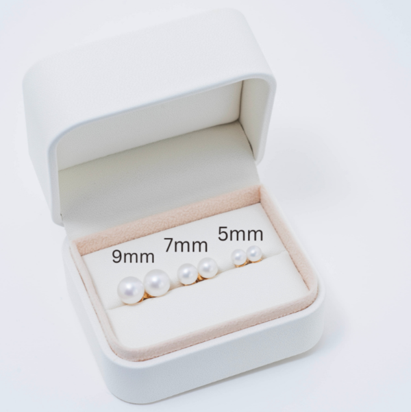 An open jewelry box showcases three pairs of freshwater pearl stud earrings, sized at 9mm, 7mm, and the Youth 5mm variant. These AAA+ quality pearls are arranged from left to right on a beige cushion and are beautifully set in 14k yellow gold.