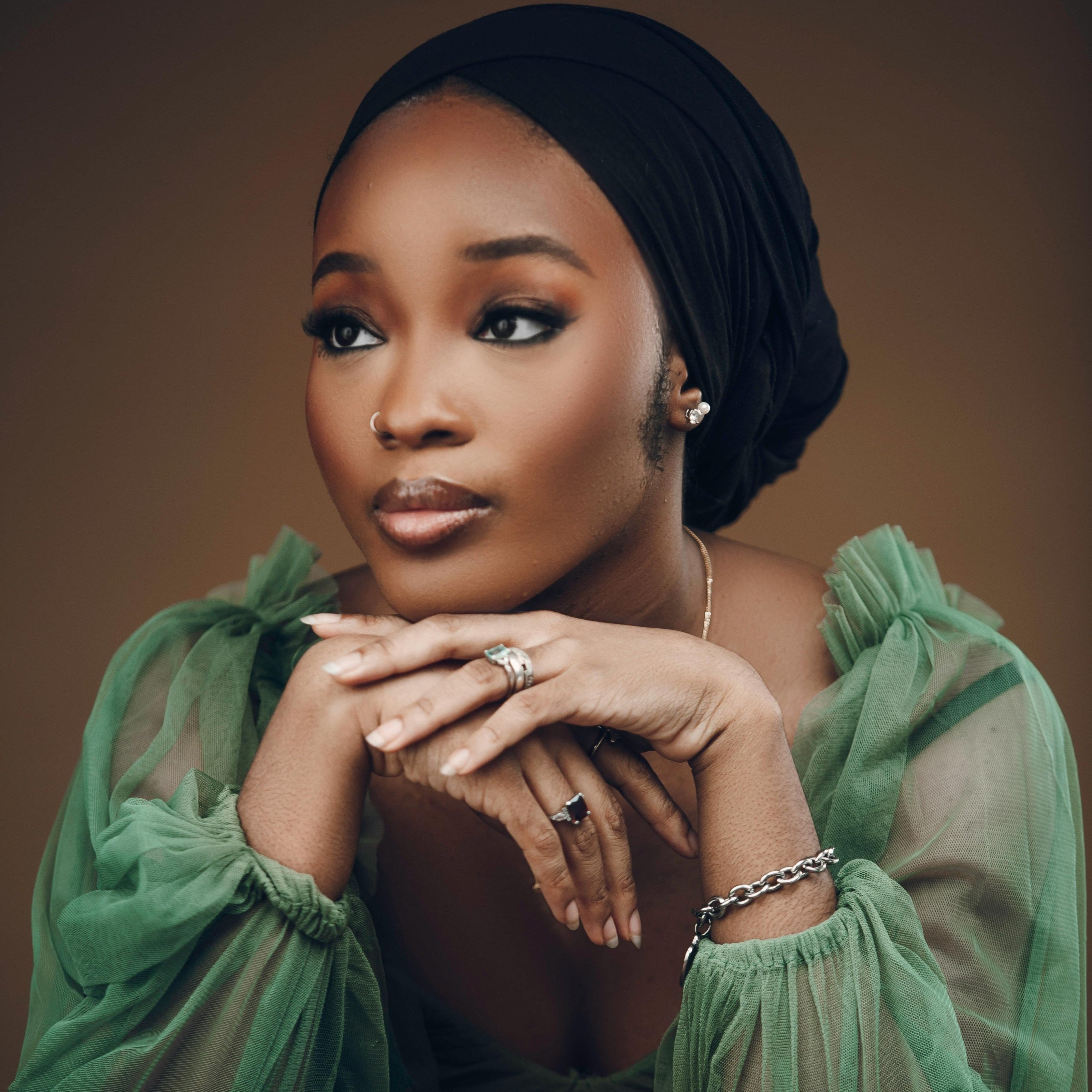 A person with a thoughtful expression rests their chin on their hands. They are wearing a sheer green top and a black headscarf, adorned with Goddess Jewelry—rings, a bracelet, and earrings. The background is a soft brown.