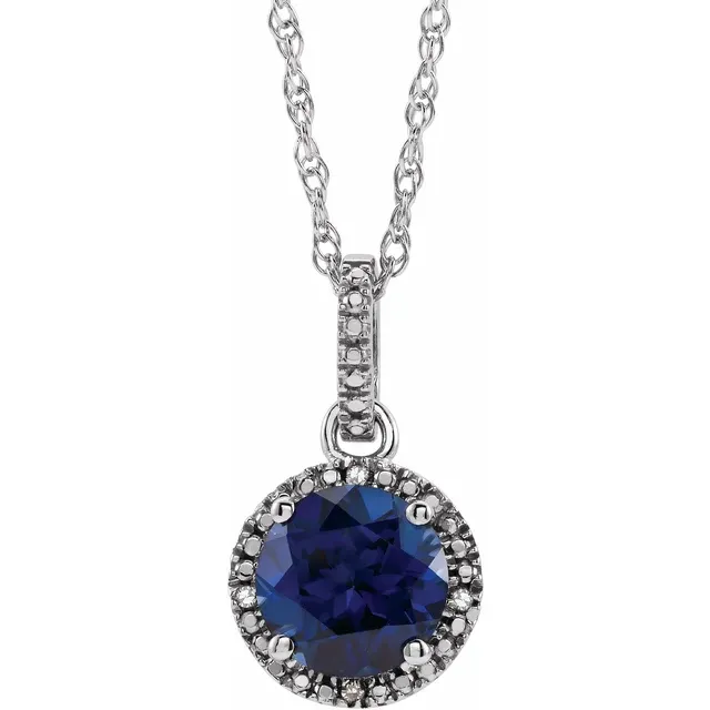 The Stuller Sterling Silver Lab-Grown Blue Sapphire Round Necklace is displayed against a white background, featuring a pendant encircled by small white stones. The intricately designed chain enhances its elegant presentation as a piece of fine jewelry.