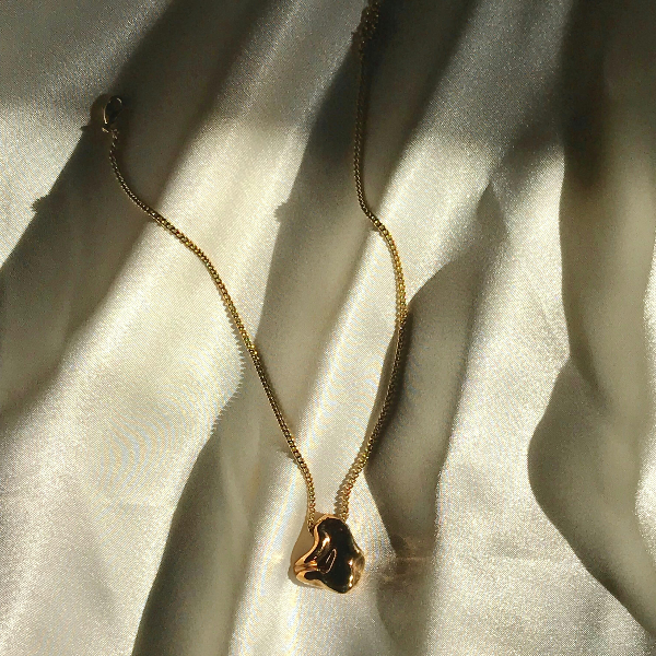 A gold necklace with an abstract, wavy pendant from Goddess Jewelry rests on white, textured fabric. The light casts soft shadows, highlighting the pendant's smooth, reflective surface and the delicate chain.
